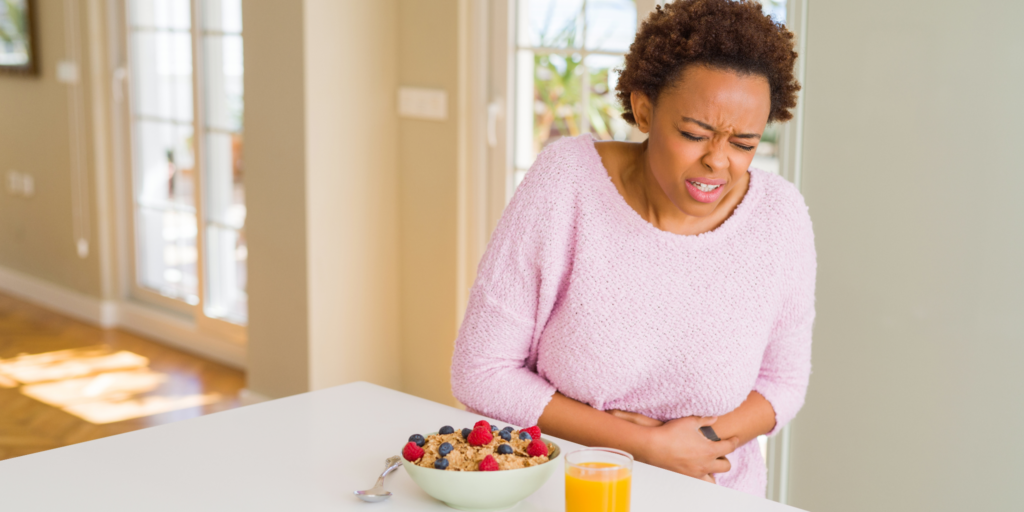 A young Black woman in a pink sweater clutching her stomach in pain due to gut irritation caused by flavored yogurt with fruit and orange juice on the counter.