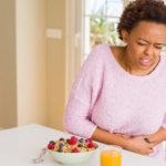 A young Black woman in a pink sweater clutching her stomach in pain due to gut irritation caused by flavored yogurt with fruit and orange juice on the counter.