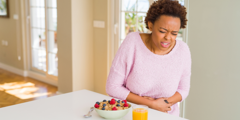 A young Black woman in a pink sweater clutching her stomach in pain due to gut irritation caused by flavored yogurt with fruit and orange juice on the counter.