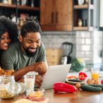Young Black couple leaning over kitchen counter, looking for cheap magnesium foods online using a laptop.