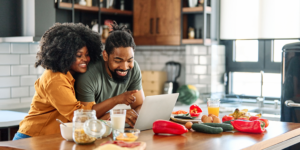 Young Black couple leaning over kitchen counter, looking for cheap magnesium foods online using a laptop.