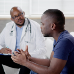 A Black man in a navy blue t-shirt sits across from a Black male doctor in a white coat and stethoscope, speaking expressively with his hands during a medical consultation about borderline PSA results.