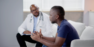 A Black man in a navy blue t-shirt sits across from a Black male doctor in a white coat and stethoscope, speaking expressively with his hands during a medical consultation about borderline PSA results.