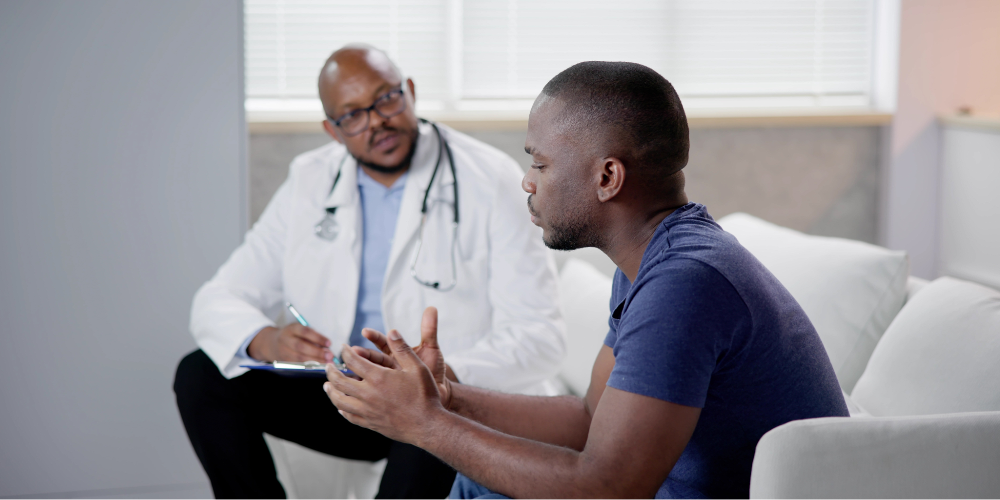 A Black man in a navy blue t-shirt sits across from a Black male doctor in a white coat and stethoscope, speaking expressively with his hands during a medical consultation about borderline PSA results.