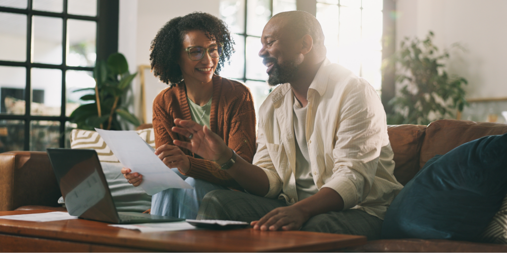 A Black couple sitting together on a brown leather couch in a bright living room, smiling as they look over at-home cancer screening kit paperwork and a laptop. The woman holds a document while the man gestures toward the screen.