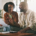 A Black couple sitting together on a brown leather couch in a bright living room, smiling as they look over at-home cancer screening kit paperwork and a laptop. The woman holds a document while the man gestures toward the screen.