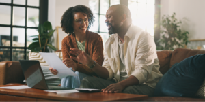 A Black couple sitting together on a brown leather couch in a bright living room, smiling as they look over at-home cancer screening kit paperwork and a laptop. The woman holds a document while the man gestures toward the screen.