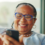A close-up shot of an older Black woman with braided hair, smiling brightly as she looks at her smartphone for heart failure apps. She wears blue-rimmed glasses, gold hoop earrings, a pearl necklace, and a light blue cardigan over a white top.