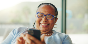 A close-up shot of an older Black woman with braided hair, smiling brightly as she looks at her smartphone for heart failure apps. She wears blue-rimmed glasses, gold hoop earrings, a pearl necklace, and a light blue cardigan over a white top.