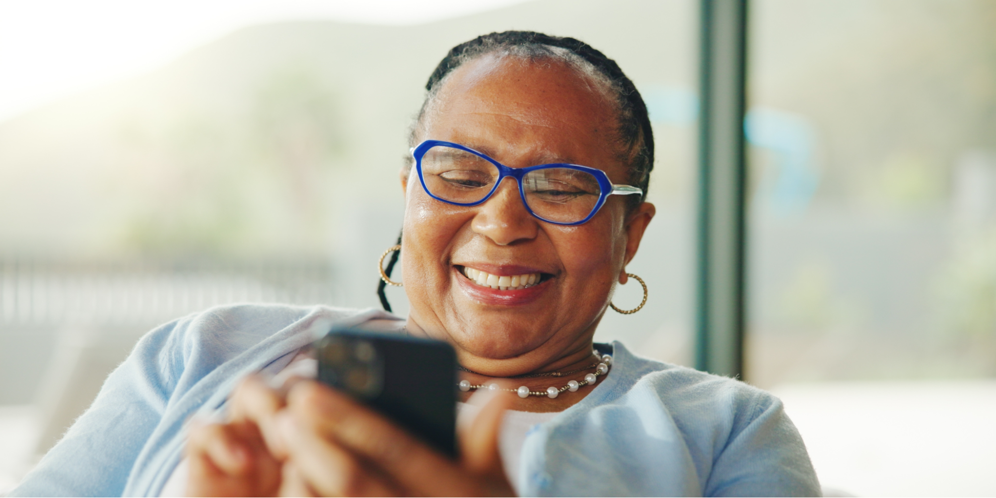 A close-up shot of an older Black woman with braided hair, smiling brightly as she looks at her smartphone for heart failure apps. She wears blue-rimmed glasses, gold hoop earrings, a pearl necklace, and a light blue cardigan over a white top.