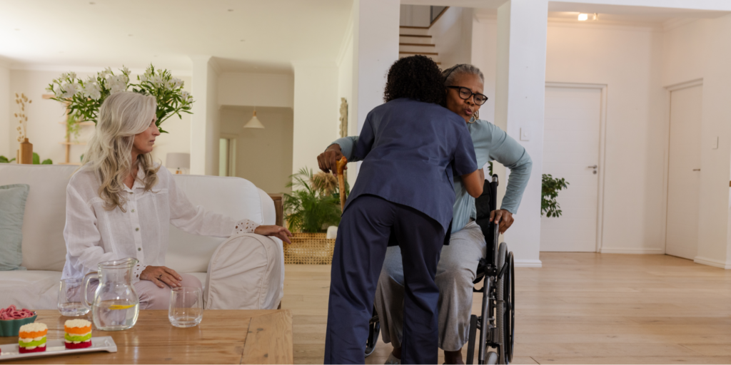 A healthcare worker in dark blue scrubs hugs an older Black woman with Alzheimer’s who is using a wheelchair and holding a wooden cane (key home safety products). In the background, a white woman with long gray hair watches supportively from a white sofa in a bright, spacious living room.