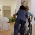 A healthcare worker in dark blue scrubs hugs an older Black woman with Alzheimer’s who is using a wheelchair and holding a wooden cane (key home safety products). In the background, a white woman with long gray hair watches supportively from a white sofa in a bright, spacious living room.