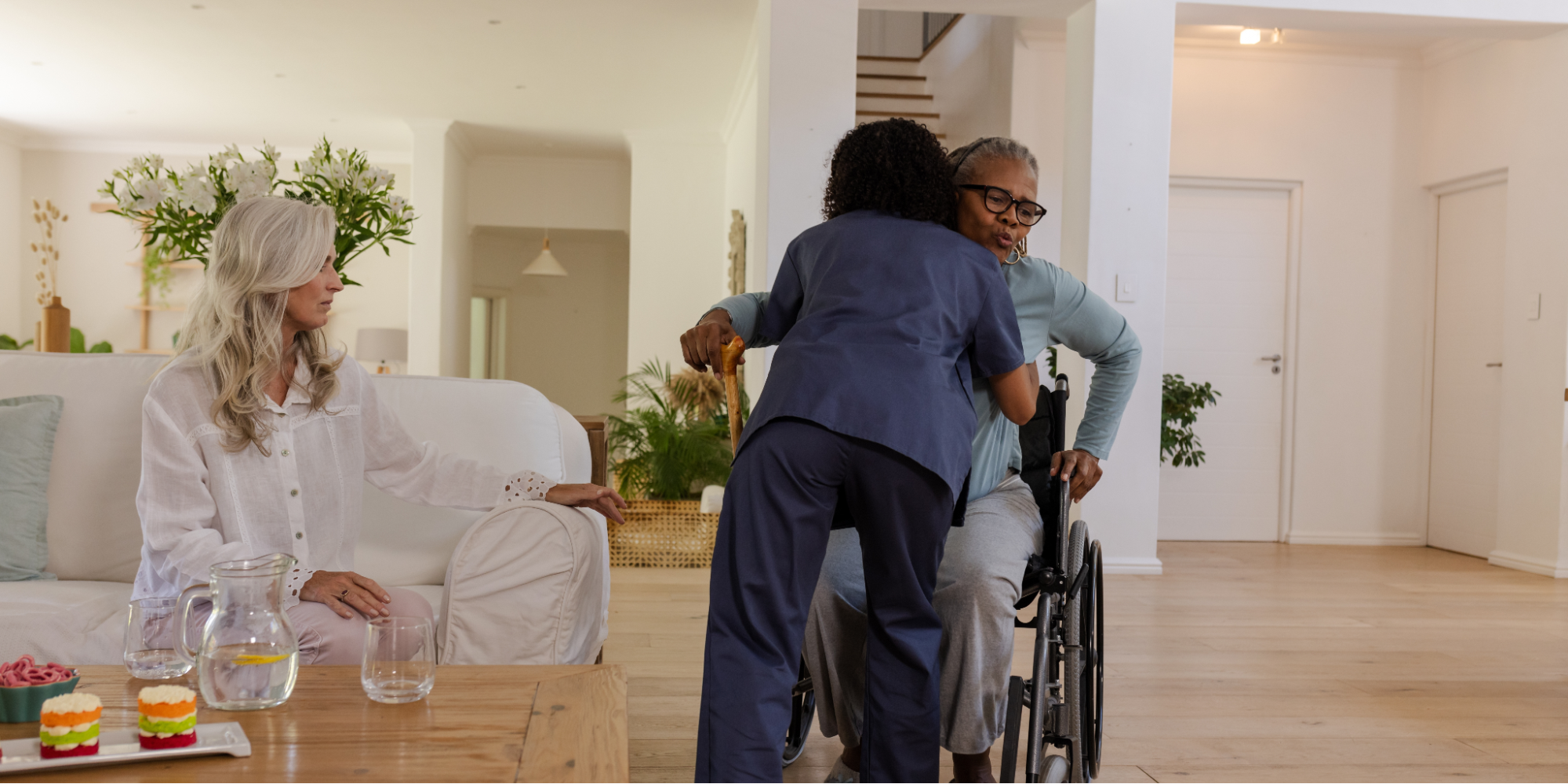 A healthcare worker in dark blue scrubs hugs an older Black woman with Alzheimer’s who is using a wheelchair and holding a wooden cane (key home safety products). In the background, a white woman with long gray hair watches supportively from a white sofa in a bright, spacious living room.