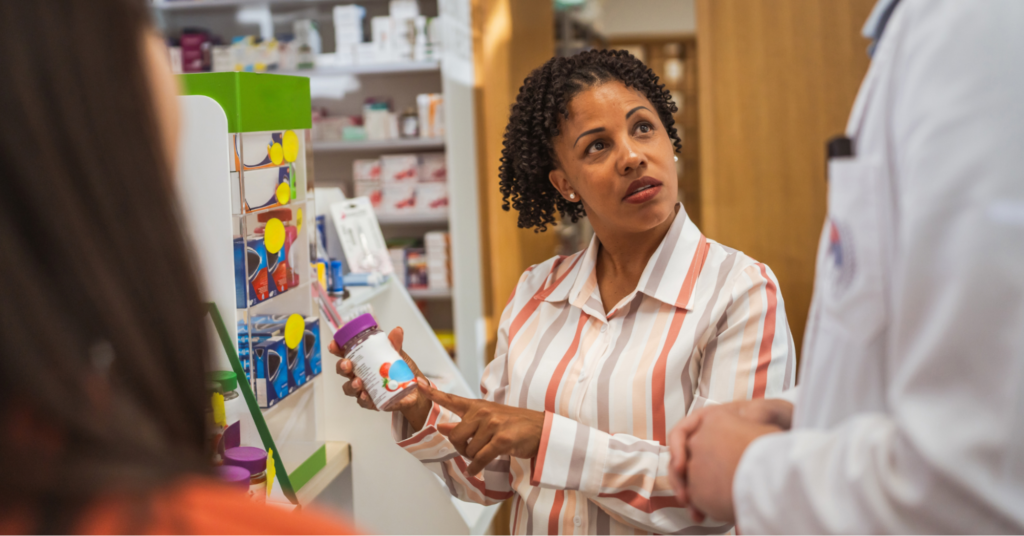 A Black woman in a striped orange and white collared shirt holding ulcerative colitis supplements in her hand as she consults with a pharmacist.