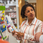 A Black woman in a striped orange and white collared shirt holding ulcerative colitis supplements in her hand as she consults with a pharmacist.