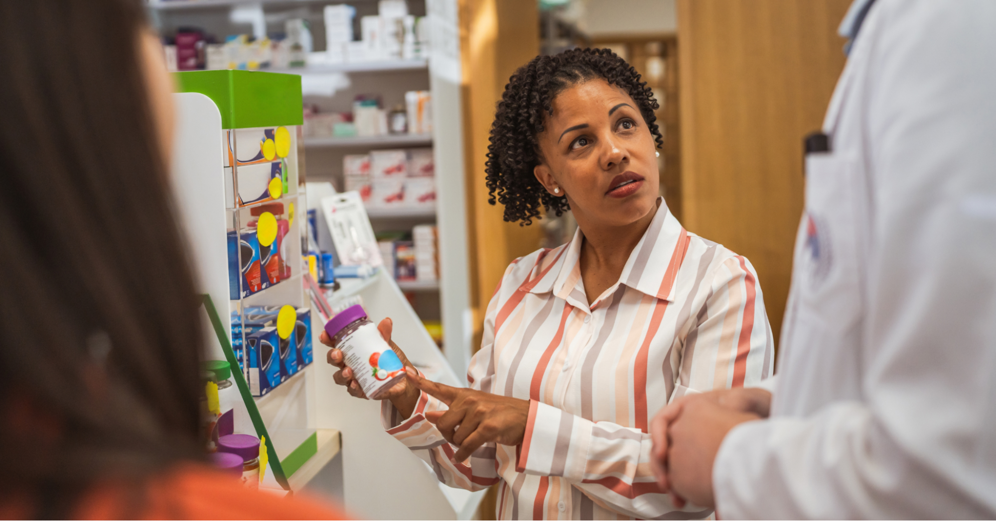 A Black woman in a striped orange and white collared shirt holding ulcerative colitis supplements in her hand as she consults with a pharmacist.