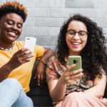 Two smiling LGBTQ+ people sit together on a dark gray sofa with decorative pillows, each happily looking at their own smartphone against a gray brick wall background. The woman on the left has short hair and a yellow top, while the woman on the right has long curly hair and glasses.