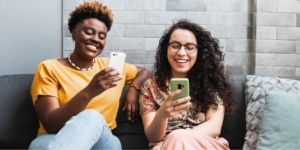 Two smiling LGBTQ+ people sit together on a dark gray sofa with decorative pillows, each happily looking at their own smartphone against a gray brick wall background. The woman on the left has short hair and a yellow top, while the woman on the right has long curly hair and glasses.