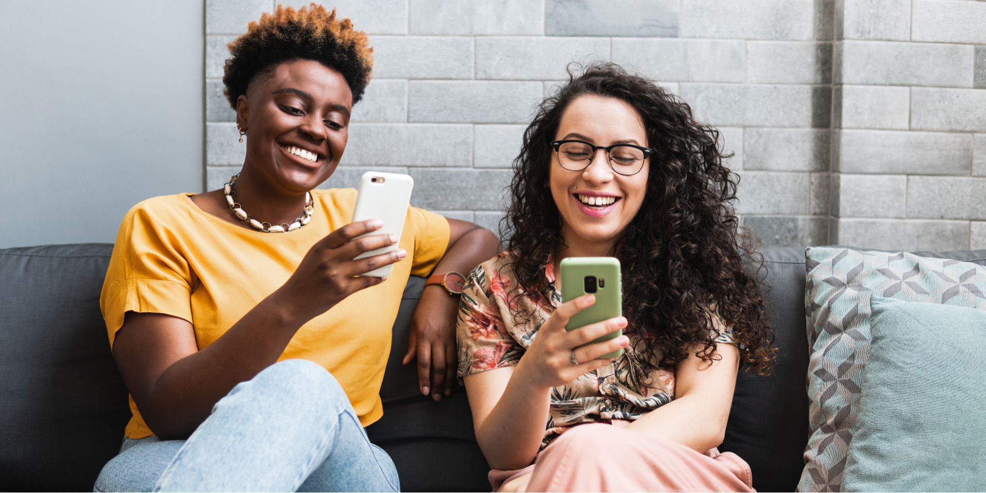 Two smiling LGBTQ+ people sit together on a dark gray sofa with decorative pillows, each happily looking at their own smartphone against a gray brick wall background. The woman on the left has short hair and a yellow top, while the woman on the right has long curly hair and glasses.