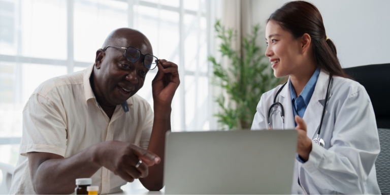 A smiling female Asian doctor in a white lab coat and stethoscope explaining colonoscopy prep information on a laptop screen to an older Black male patient. The patient lifts his glasses and points at the screen during the consultation.