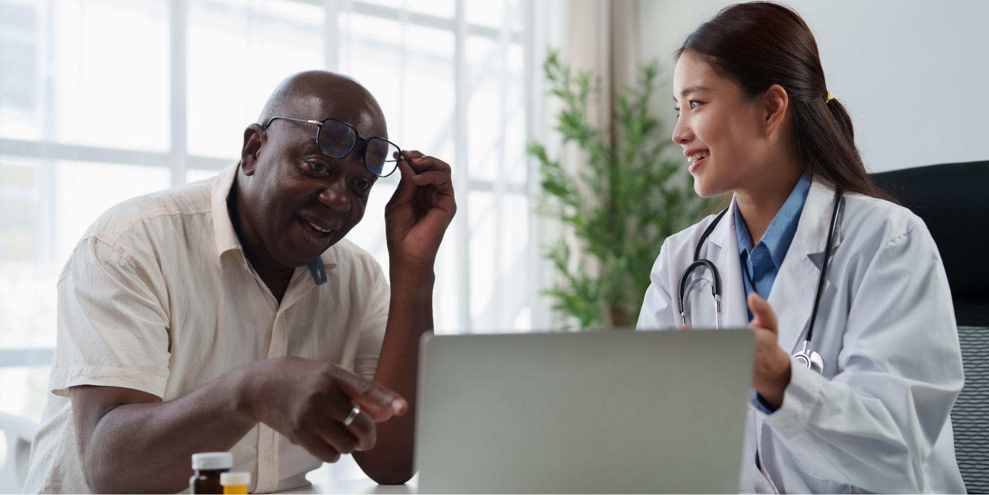 A smiling female Asian doctor in a white lab coat and stethoscope explaining colonoscopy prep information on a laptop screen to an older Black male patient. The patient lifts his glasses and points at the screen during the consultation.