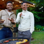 Two young Black men grilling a heart-healthy dinner at a cookout outdoors with wine in their hands.