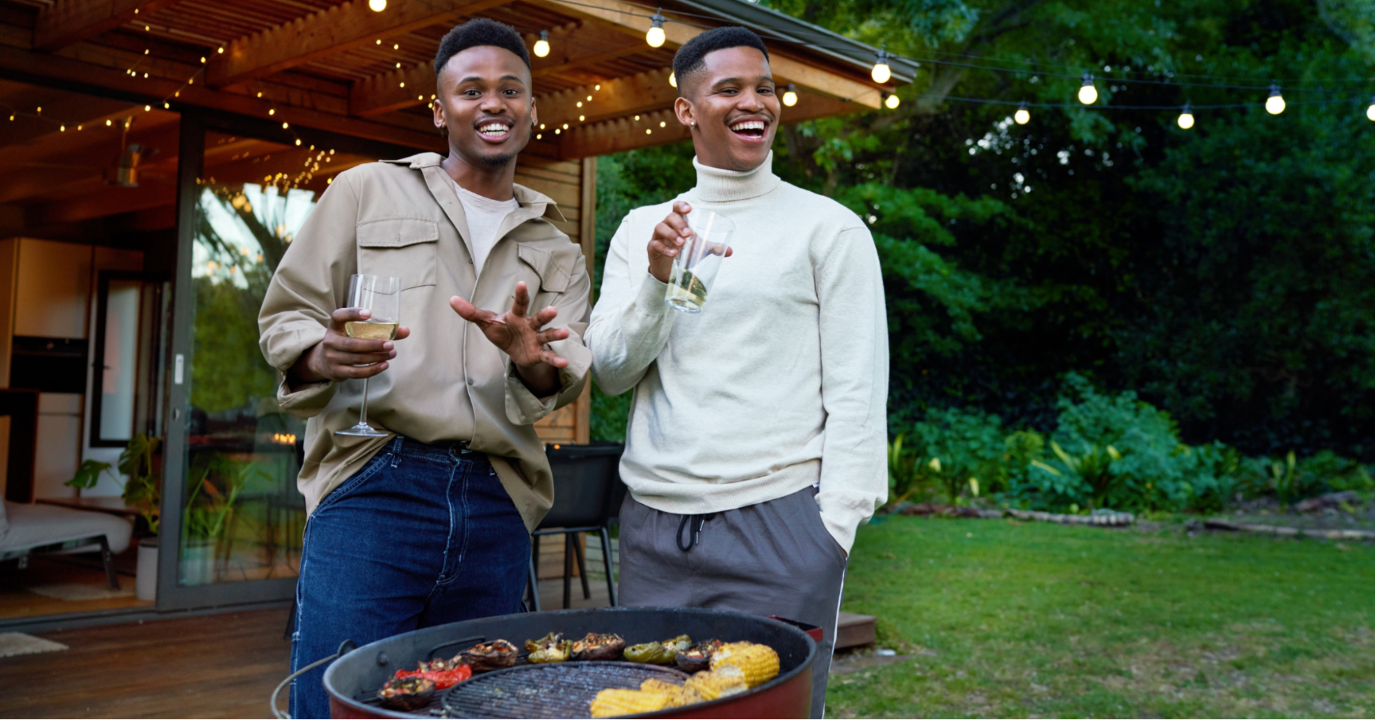 Two young Black men grilling a heart-healthy dinner at a cookout outdoors with wine in their hands.