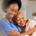 Black nurse hugging an elderly Black woman on the couch in a memory care facility.