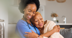 Black nurse hugging an elderly Black woman on the couch in a memory care facility.