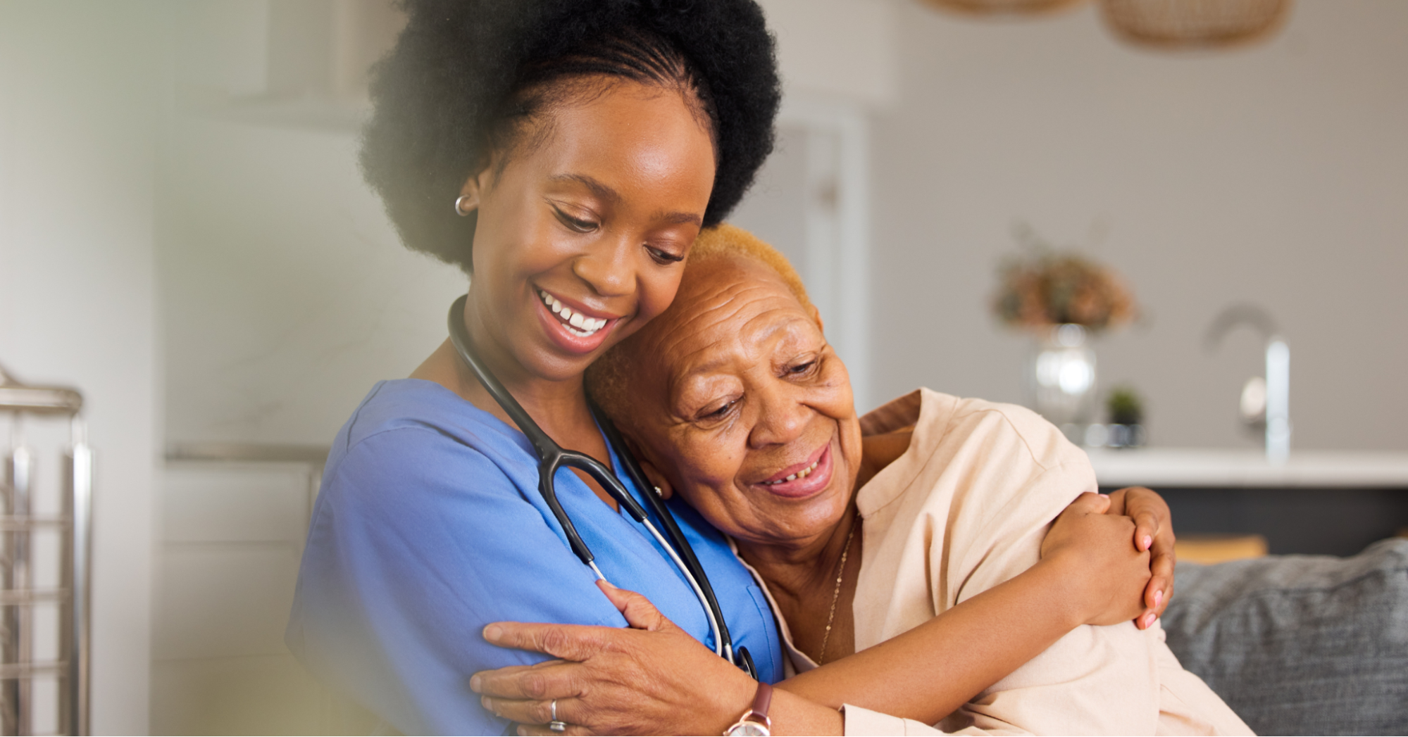 Black nurse hugging an elderly Black woman on the couch in a memory care facility.