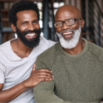 Elderly Black father with beard and glasses sitting with middle-aged Black son on the couch, smiling at the camera after having a conversation about prostate screening
