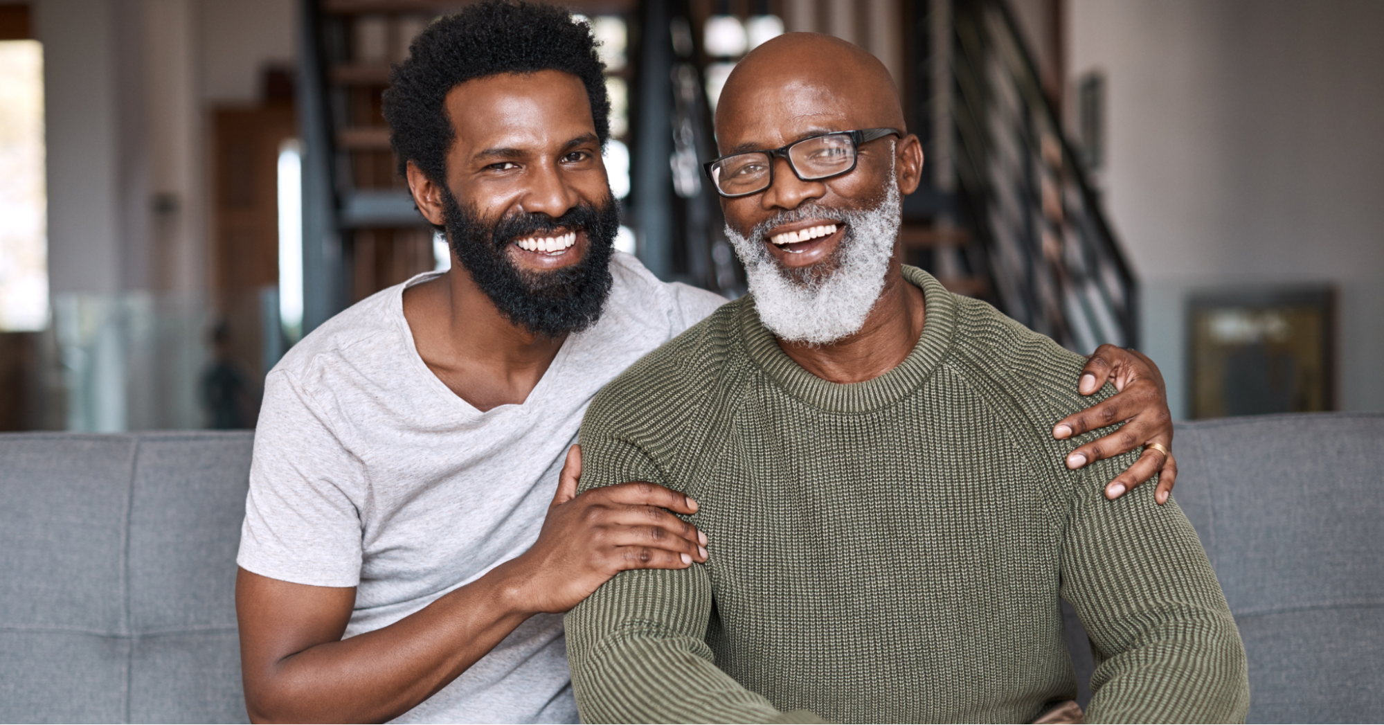 Elderly Black father with beard and glasses sitting with middle-aged Black son on the couch, smiling at the camera after having a conversation about prostate screening
