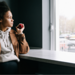 Young Black woman in brown jacket eating an apple and looking at the window, wondering if you can go in the sun if you have lupus.
