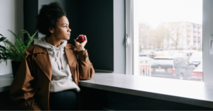 Young Black woman in brown jacket eating an apple and looking at the window, wondering if you can go in the sun if you have lupus.