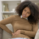 Young Black woman sitting on the couch in pain holding her right side due to back pain, one of the lung cancer symptoms in women