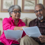 Older hetero Black couple sitting on a beige couch, wearing glasses and reviewing documents, such as prostate cancer screening guidelines