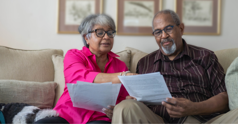 Older hetero Black couple sitting on a beige couch, wearing glasses and reviewing documents, such as prostate cancer screening guidelines