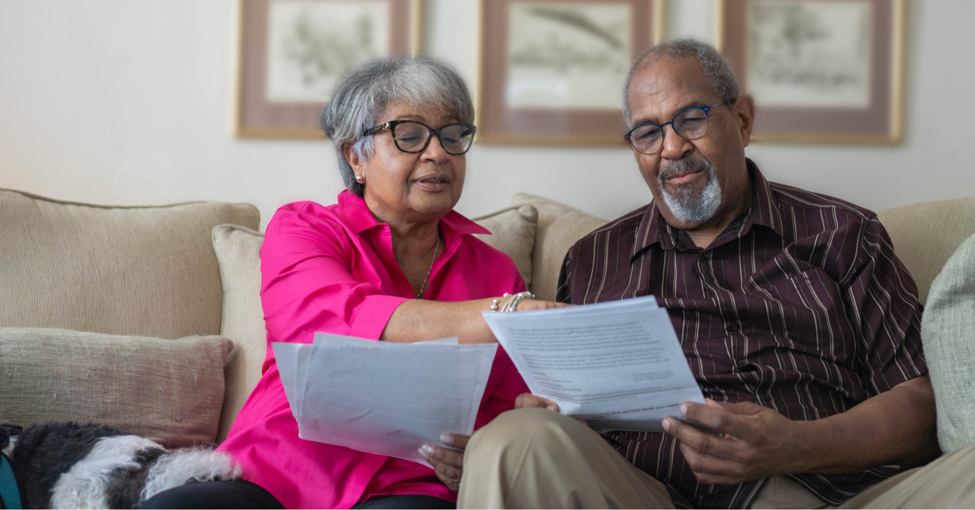 Older hetero Black couple sitting on a beige couch, wearing glasses and reviewing documents, such as prostate cancer screening guidelines