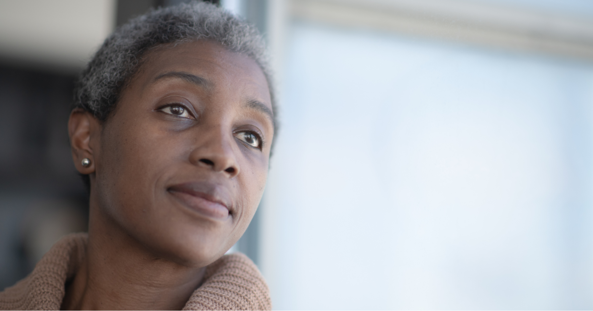 Middle-aged Black woman in a brown turtle neck looking out the window, wondering about breast cancer screening guidelines for herself and loved ones