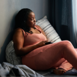 Young Black woman in a peach yoga set, sitting on the bed and looking out the window with a phone in her hand, googling her symptoms to understand the different types of lupus.