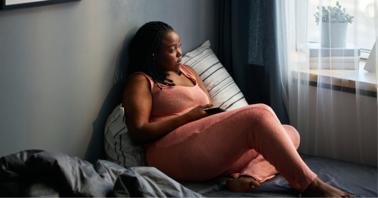 Battling Blood Cancers: Understanding the Disparities in Black Communities 2 Young Black woman in a peach yoga set, sitting on the bed and looking out the window with a phone in her hand, googling her symptoms to understand the different types of lupus.