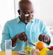 African american bald senior man wearing eyeglasses eating fruits for lunch on dining table at home
